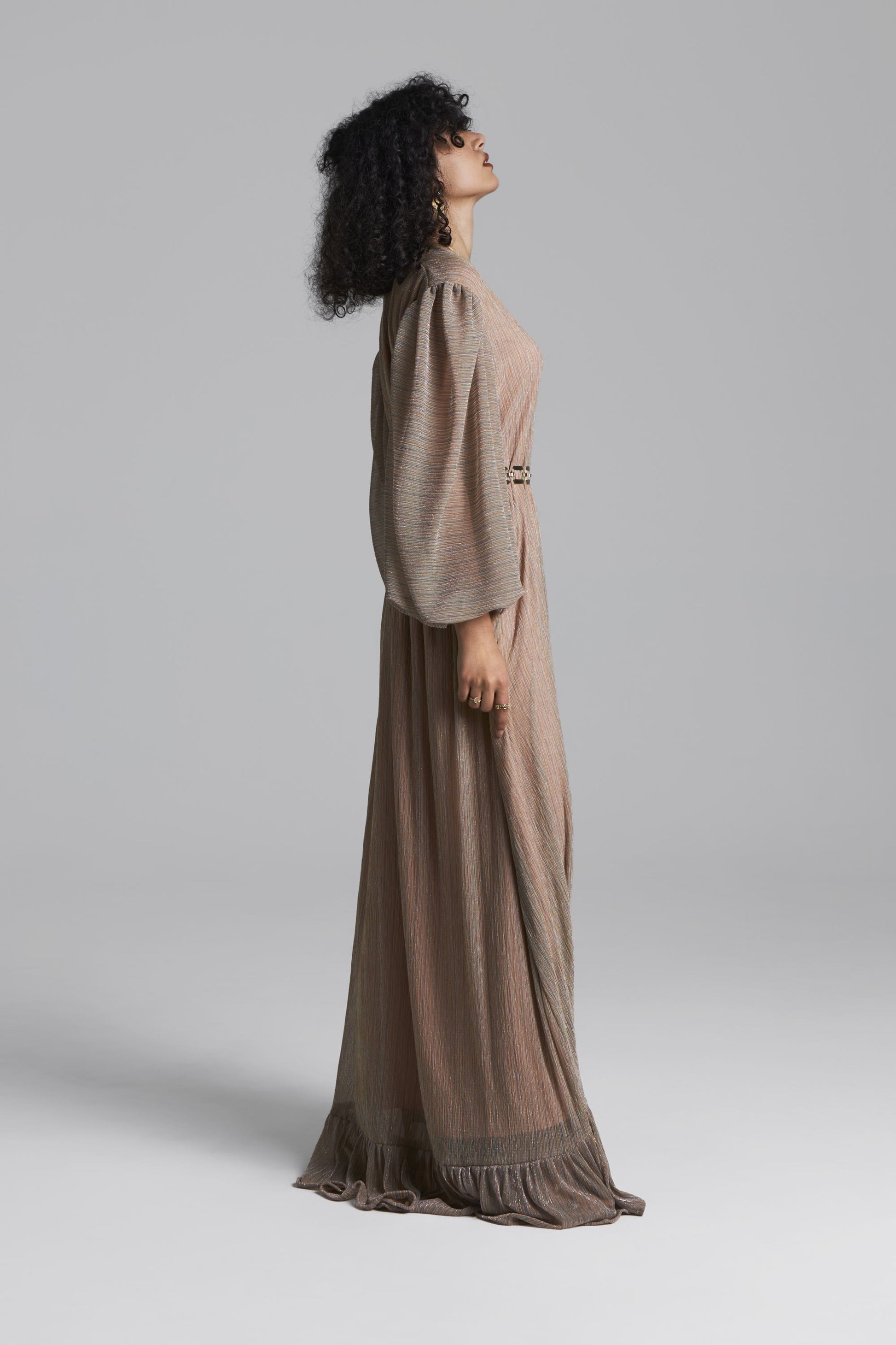 A model with curly black hair wearing a long-sleeved, floor-length taupe maxi dress with a matching belt, side view photographed against a neutral gray studio background.