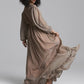 A model with curly black hair wearing a long-sleeved, floor-length taupe maxi dress with a matching belt, 3/4 side view photographed against a neutral gray studio background.