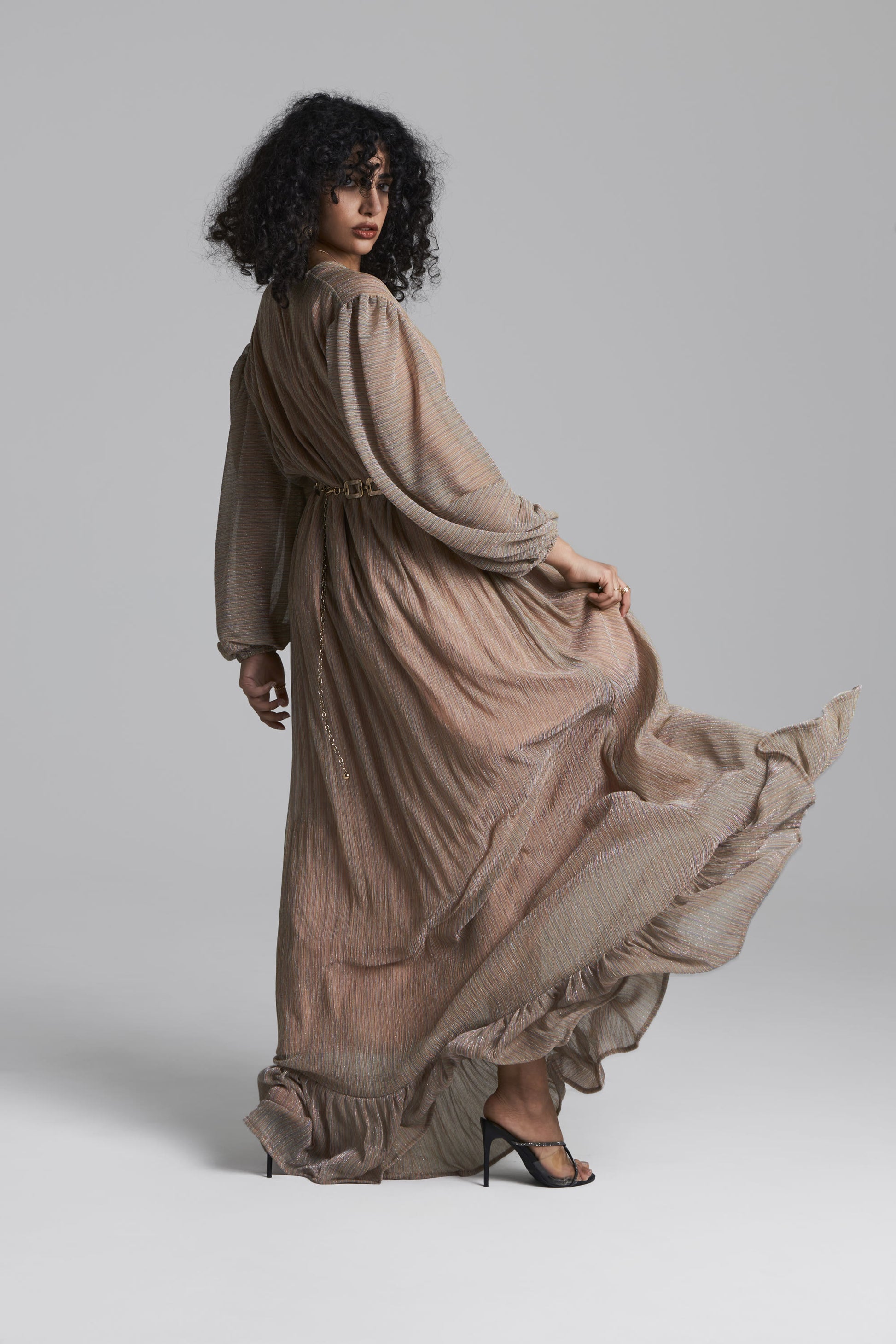 A model with curly black hair wearing a long-sleeved, floor-length taupe maxi dress with a matching belt, 3/4 side view photographed against a neutral gray studio background.