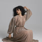 A woman with curly black hair kneels on a light gray studio floor wearing a long-sleeved, pleated taupe-brown dress with a matching belt. She is posed with one hand in her hair, wearing white hoop earrings, against a solid gray background.