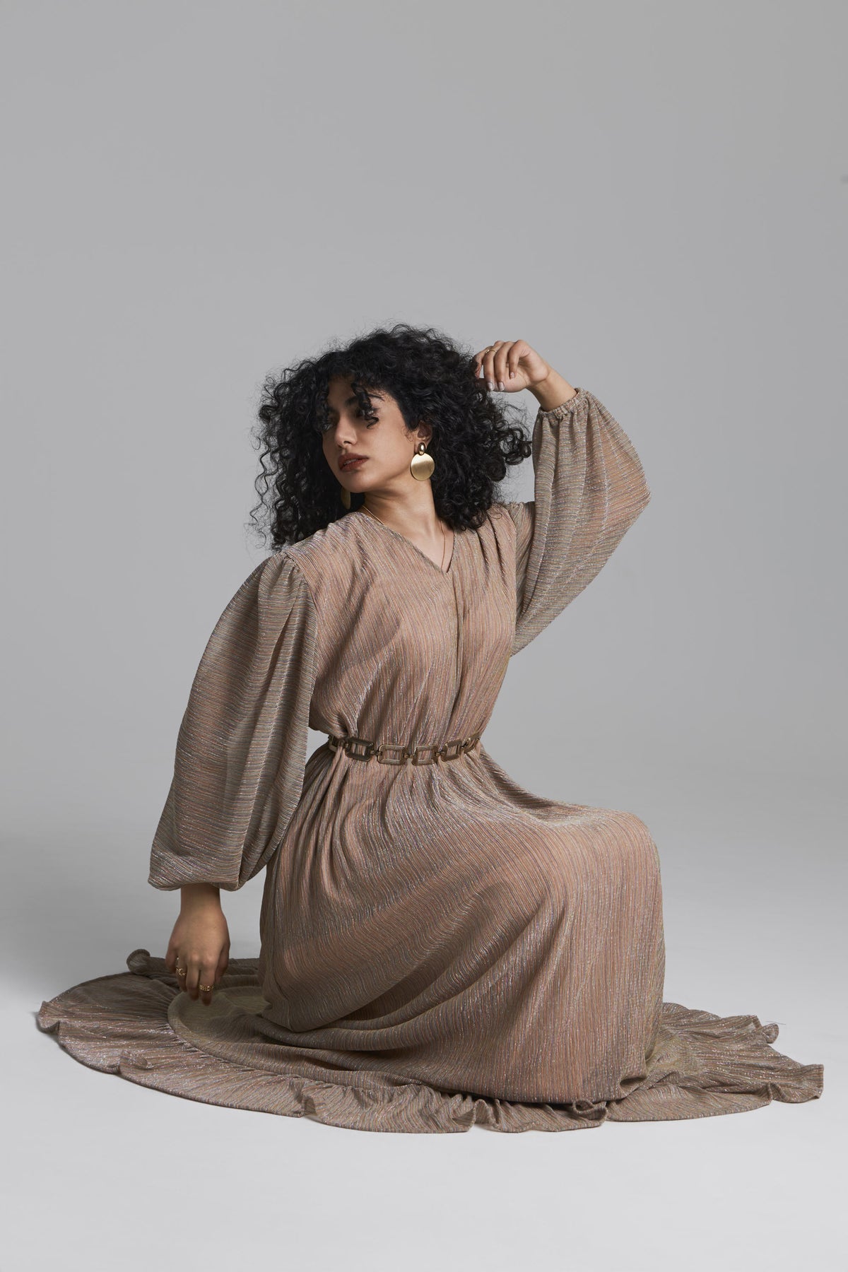 A woman with curly black hair kneels on a light gray studio floor wearing a long-sleeved, pleated taupe-brown dress with a matching belt. She is posed with one hand in her hair, wearing white hoop earrings, against a solid gray background.