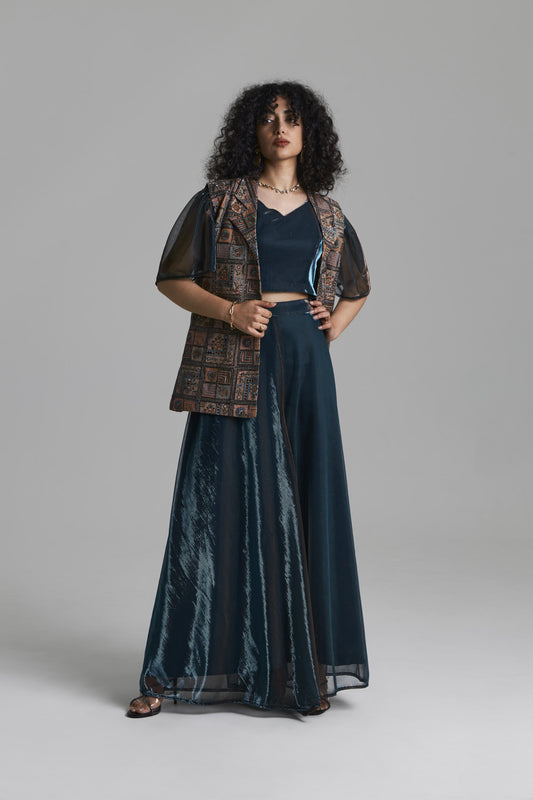 A woman modeling a dark teal two-piece outfit consisting of a sleeveless V-neck top and a matching long skirt, styled with a brown patterned vest over sheer dark sleeves, against a solid gray studio background.