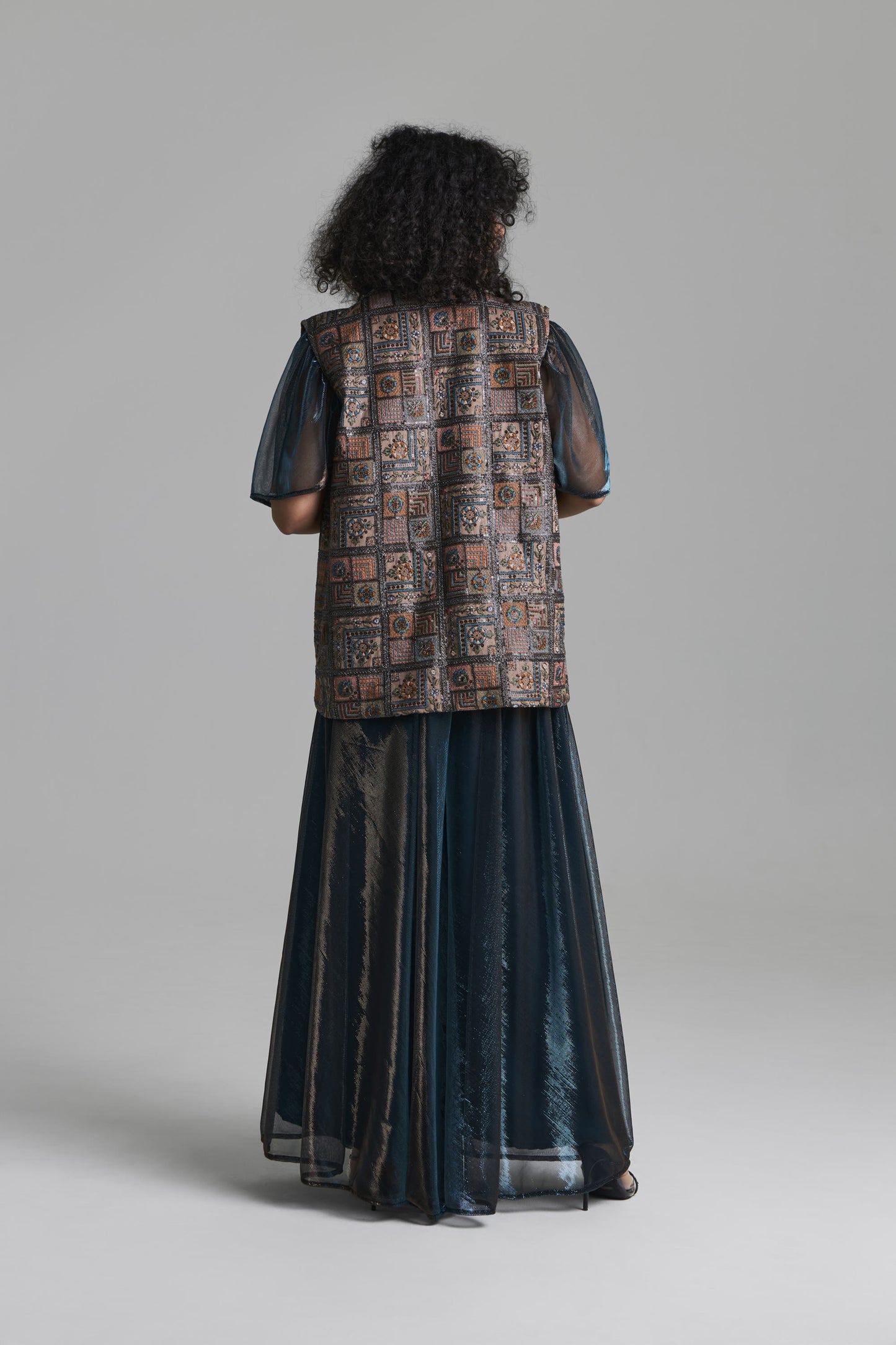 A model viewed from the back wearing a long dark teal-blue sheer skirt with a patterned brown vest layered over sheer sleeves, set against a solid gray studio background.