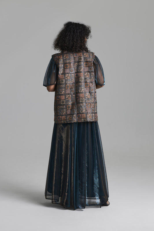 A model viewed from the back wearing a long dark teal-blue sheer skirt with a patterned brown vest layered over sheer sleeves, set against a solid gray studio background.