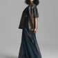 A model with curly dark hair is shown from the side wearing a dark teal blue two-piece outfit consisting of a short-sleeve crop top and a long skirt, both with a sheer patterned overlay in brown and gold tones, set against a plain gray studio background.
