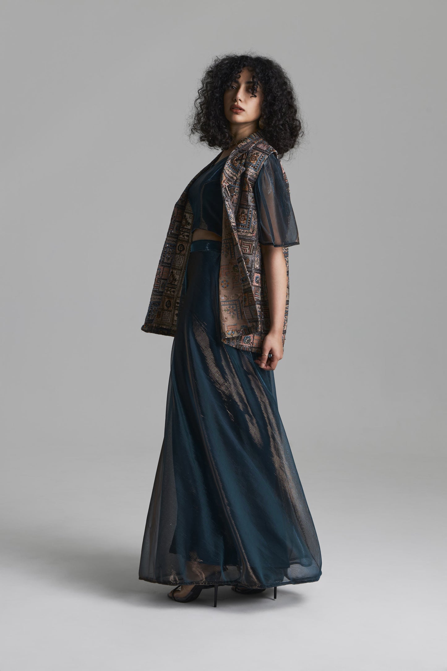 A model with curly dark hair is shown from the side wearing a dark teal blue two-piece outfit consisting of a short-sleeve crop top and a long skirt, both with a sheer patterned overlay in brown and gold tones, set against a plain gray studio background.
