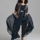 A model with curly black hair poses in a dark navy blue outfit featuring a patterned vest, a matching crop top, and wide-leg sheer trousers that are being held out to the sides. She is wearing metallic brown high-heeled sandals against a solid light gray studio background.
