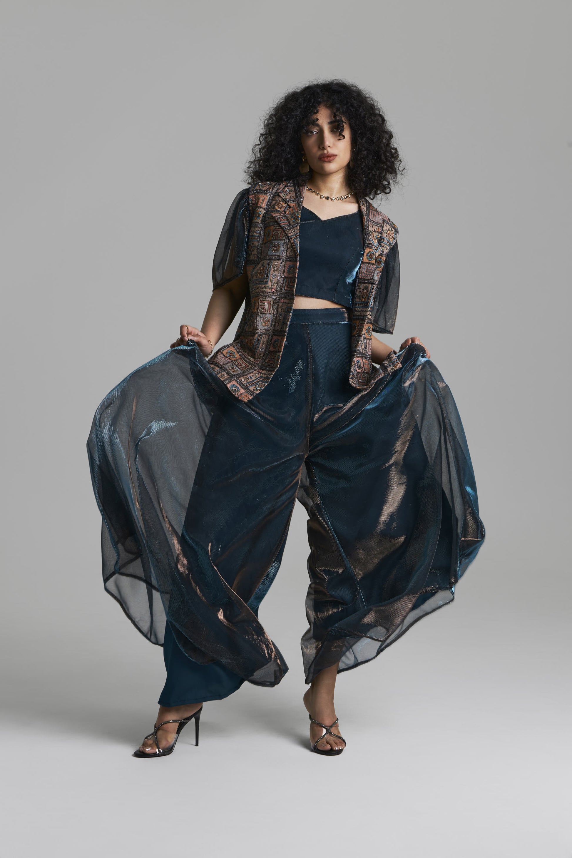 A model with curly black hair poses in a dark navy blue outfit featuring a patterned vest, a matching crop top, and wide-leg sheer trousers that are being held out to the sides. She is wearing metallic brown high-heeled sandals against a solid light gray studio background.