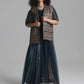 A model stands against a neutral gray studio background wearing a long dark teal sheer maxi dress with a patterned brown vest layered over it.