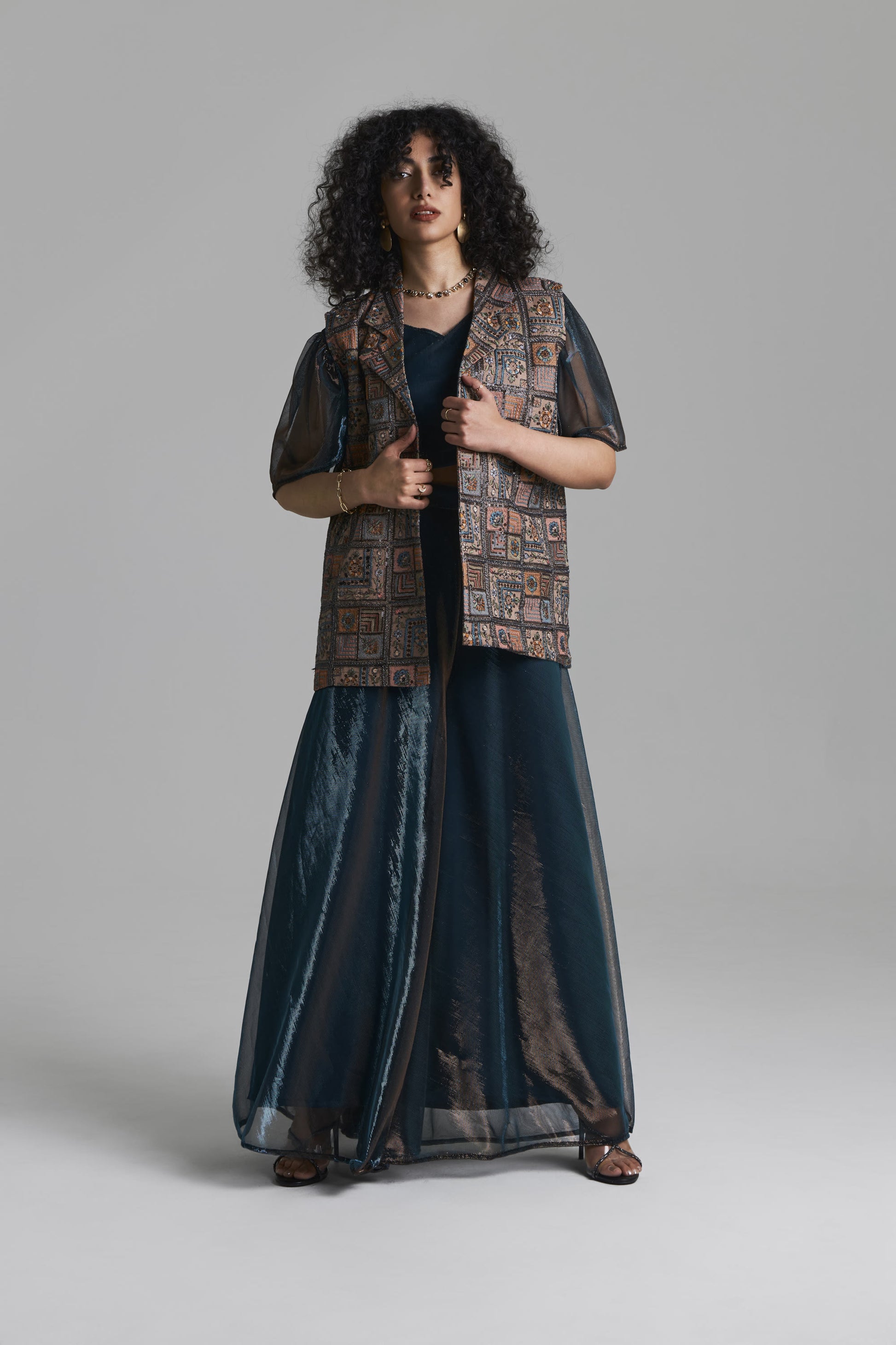 A model stands against a neutral gray studio background wearing a long dark teal sheer maxi dress with a patterned brown vest layered over it.
