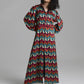 A model with curly black hair wearing a long-sleeved maxi shirt dress with a colorful zigzag chevron pattern in red, teal, yellow, and white, standing against a neutral gray studio background.