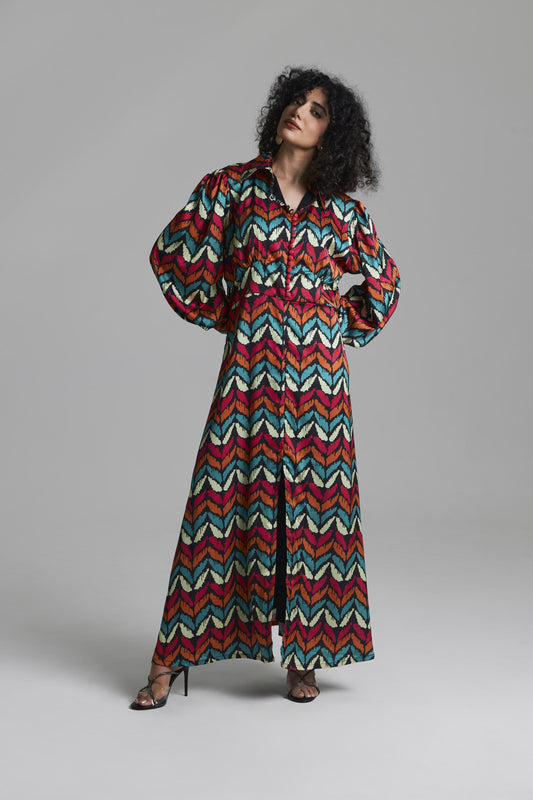 A model with curly black hair wearing a long-sleeved maxi shirt dress with a colorful zigzag chevron pattern in red, teal, yellow, and white, standing against a neutral gray studio background.