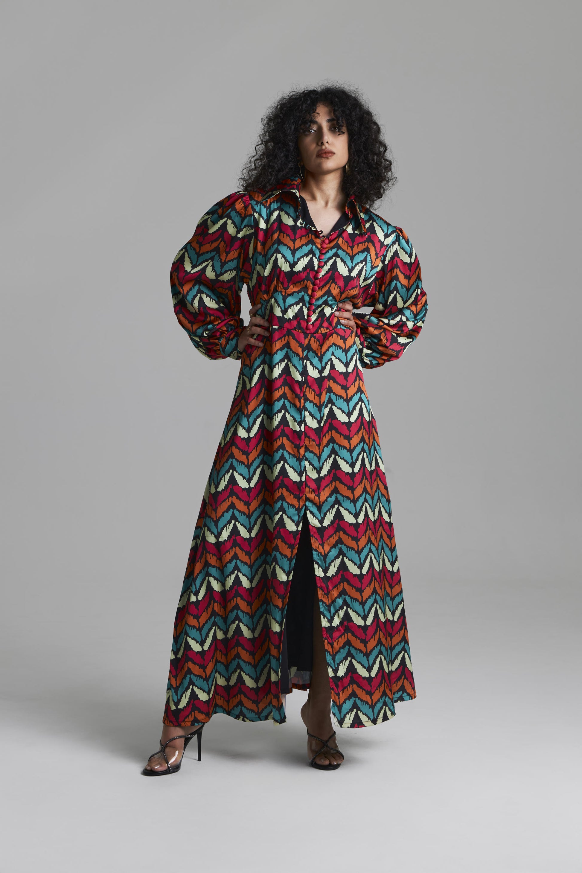 A model wearing a long-sleeved, collared maxi dress with a vibrant multicolor chevron pattern in red, teal, cream, and brown, standing against a neutral gray studio background.