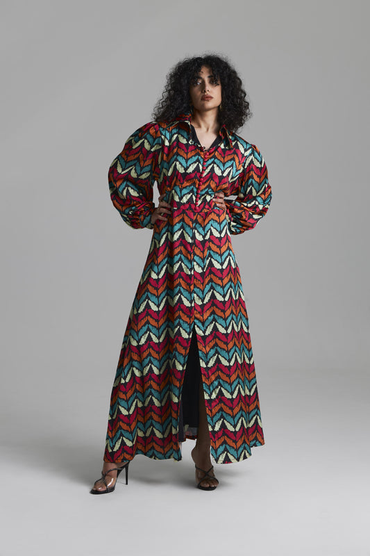 A model wearing a long-sleeved, collared maxi dress with a vibrant multicolor chevron pattern in red, teal, cream, and brown, standing against a neutral gray studio background.