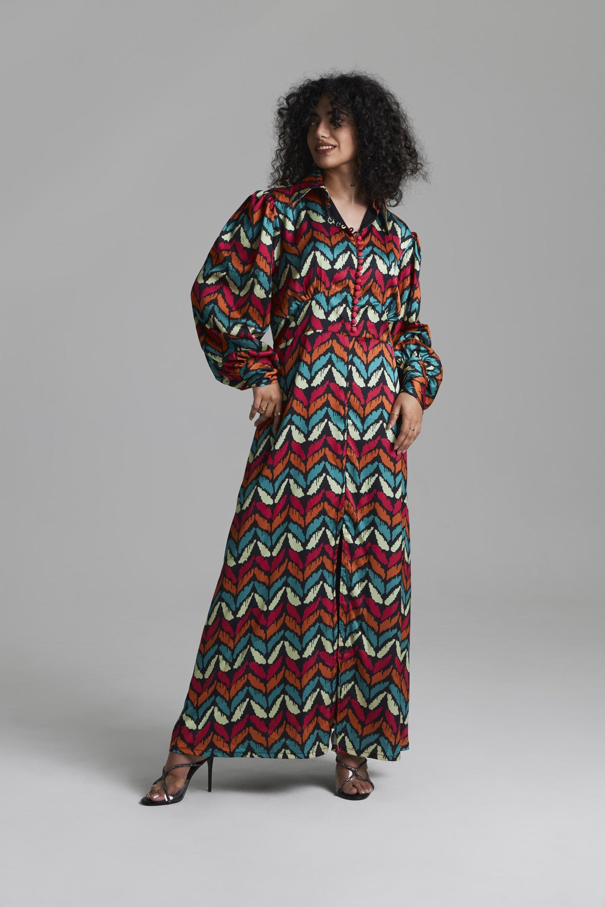 A woman modeling a long-sleeved maxi dress with a vibrant multicolor chevron pattern in red, teal, green, and cream, set against a plain gray studio background.
