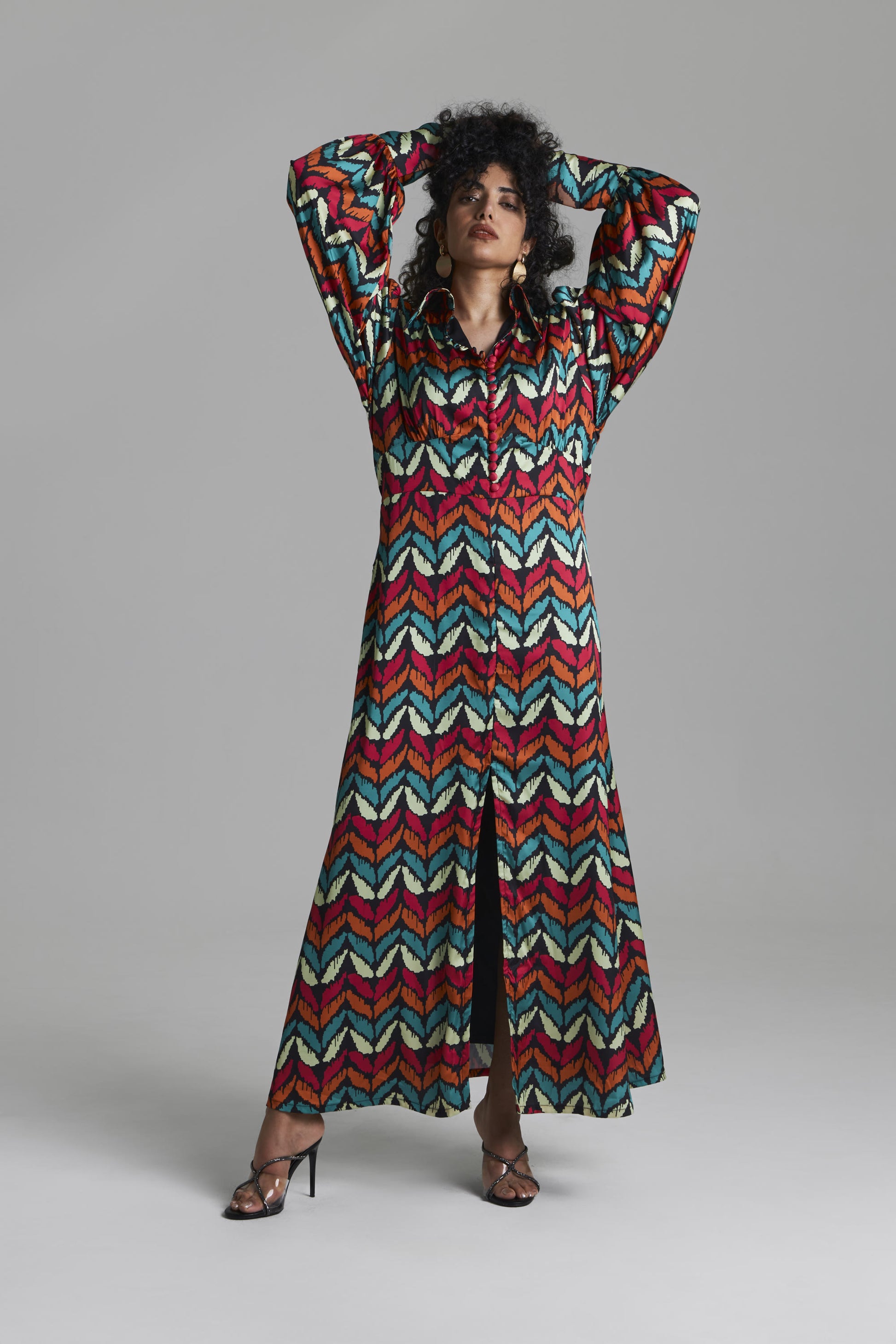 A woman modeling a long-sleeved maxi dress with a vibrant multicolor chevron pattern in red, teal, yellow, and white, set against a plain gray studio background.