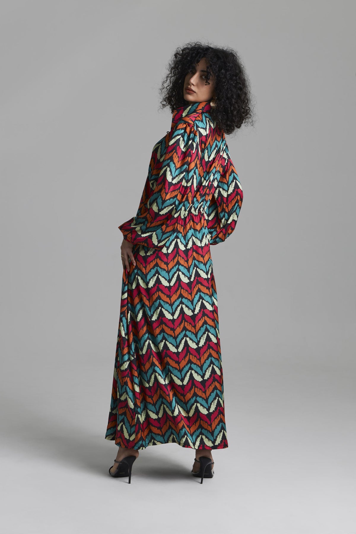 A model wearing a long-sleeved maxi dress with a vibrant multicolor chevron pattern, photographed against a neutral gray studio background.