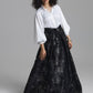 A model wearing a white long-sleeve blouse and a black high-waisted maxi skirt with a silver floral brocade pattern, standing against a neutral gray studio background.