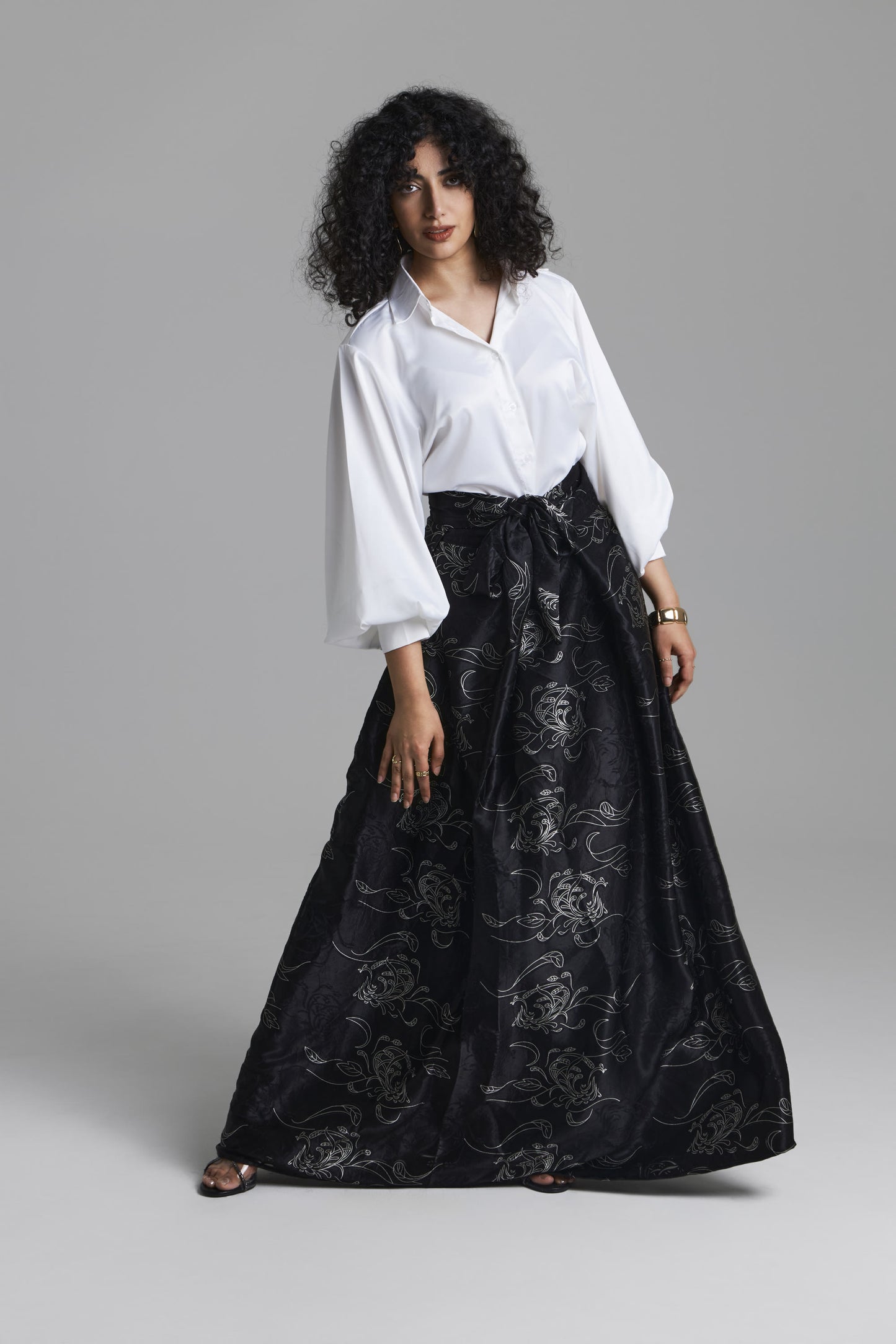 A model wearing a white long-sleeve blouse and a black high-waisted maxi skirt with a silver floral brocade pattern, standing against a neutral gray studio background.