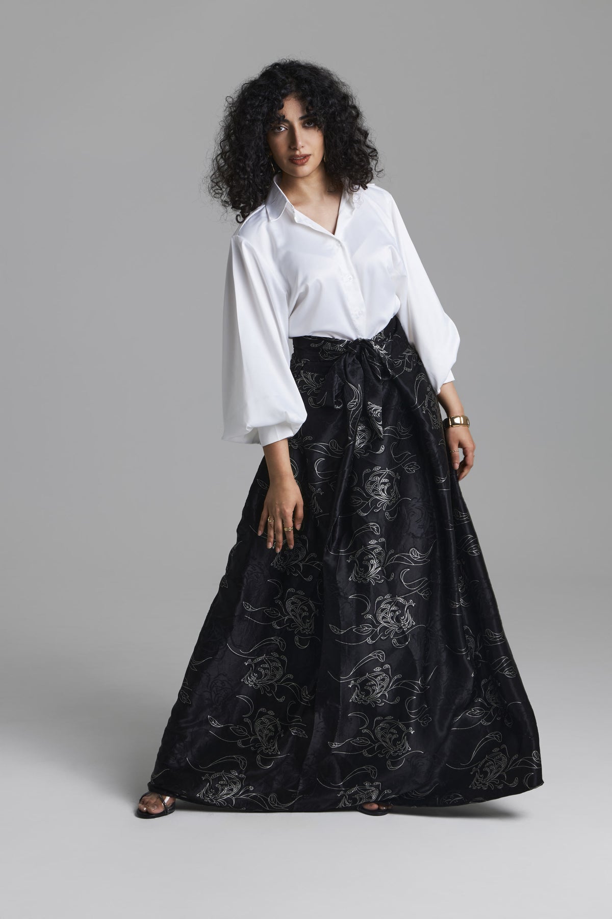 A model wearing a white long-sleeve blouse and a black high-waisted maxi skirt with a silver floral brocade pattern, standing against a neutral gray studio background.