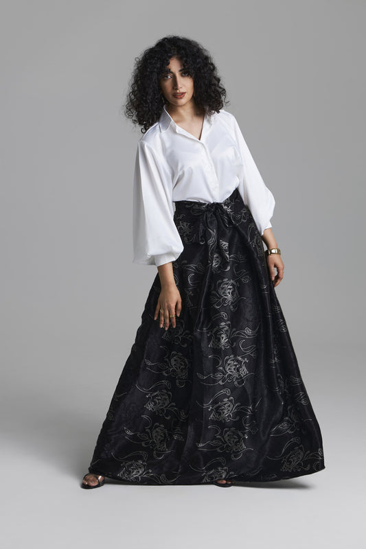 A model wearing a white long-sleeve blouse and a black high-waisted maxi skirt with a silver floral brocade pattern, standing against a neutral gray studio background.