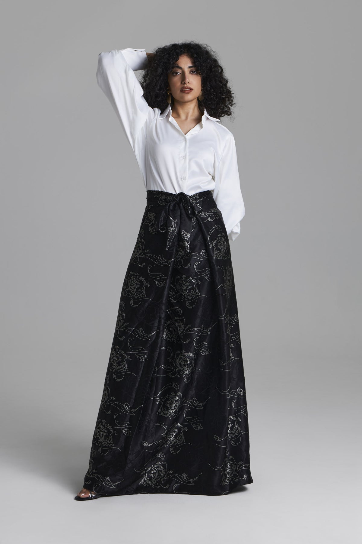 A model wearing a white long-sleeve button-up shirt and a high-waisted black maxi skirt with a silver floral jacquard pattern, posed against a solid gray studio background.