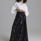 A model wearing a white long-sleeve button-up shirt and a black maxi skirt with a silver floral jacquard pattern, standing against a solid gray studio background.