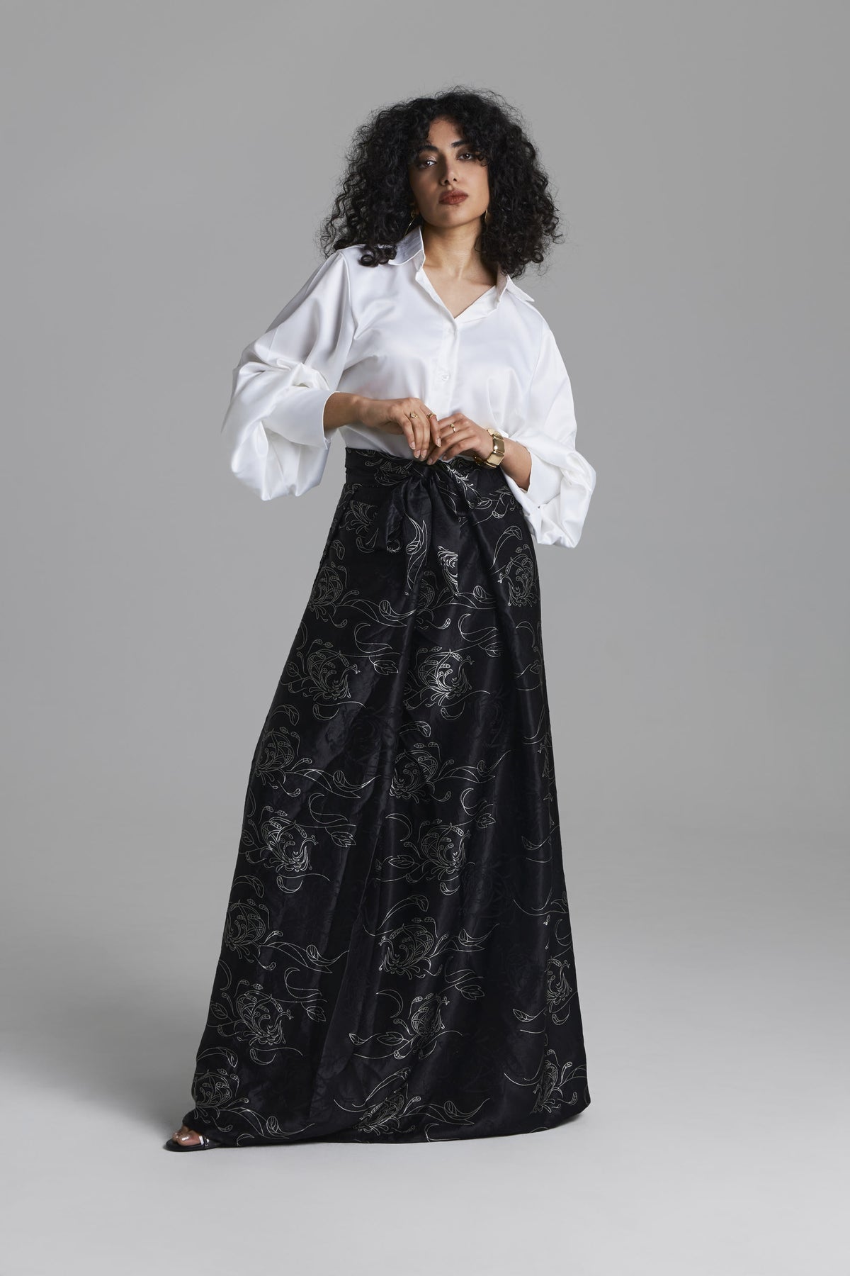 A model wearing a white long-sleeve button-up shirt and a black maxi skirt with a silver floral jacquard pattern, standing against a solid gray studio background.