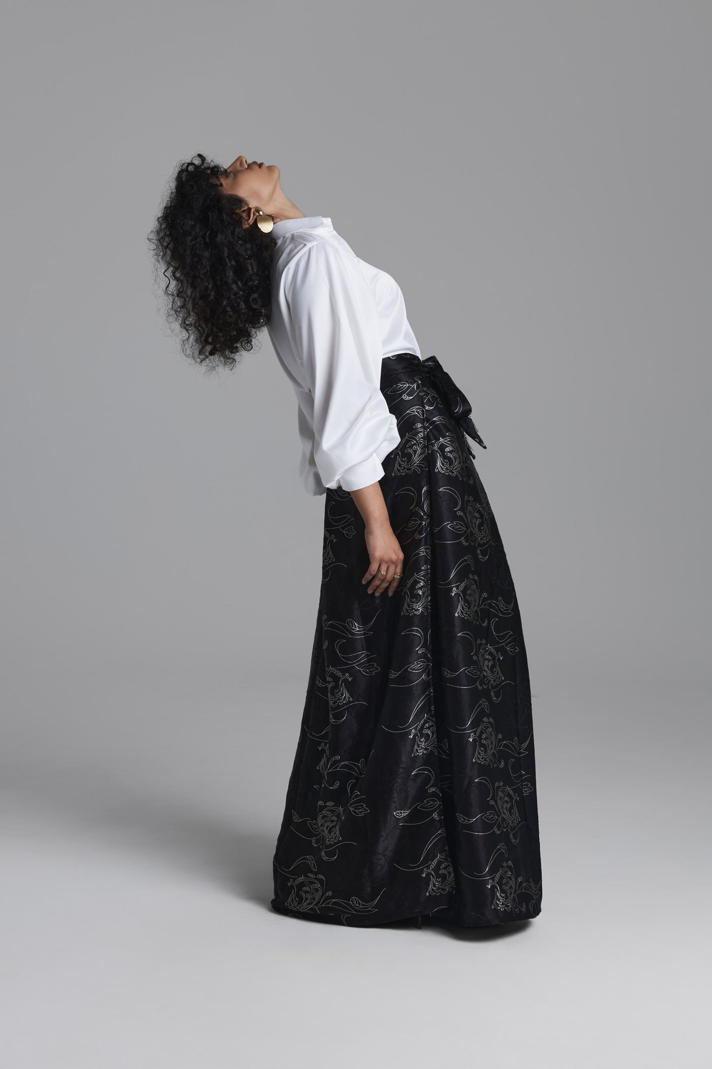A model wearing a long black skirt with a silver floral jacquard pattern and a white blouse, posing in profile against a solid gray studio background.