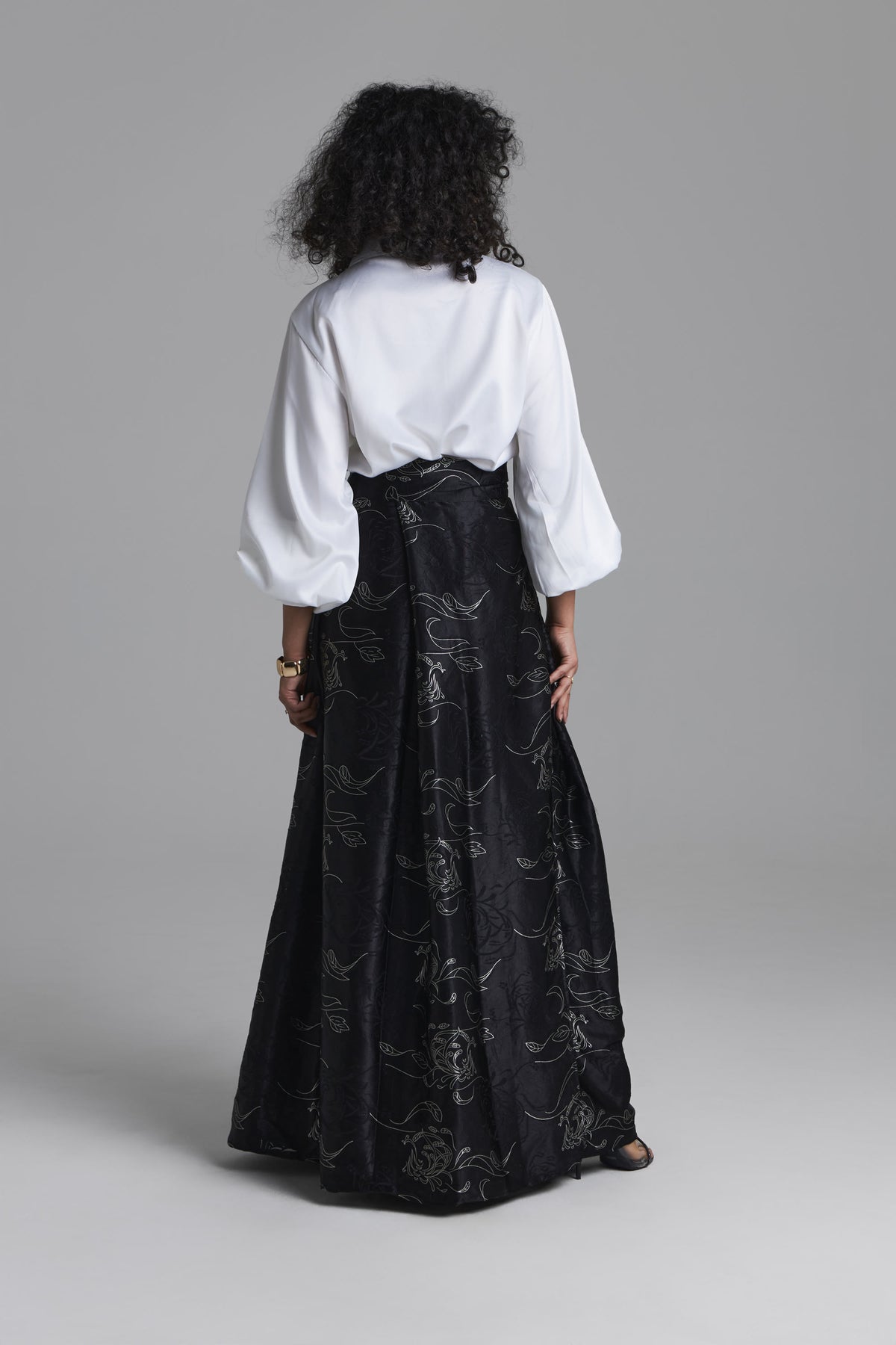A model shown from the back wearing a white blouse with voluminous three-quarter sleeves and a long black skirt featuring a subtle tonal floral pattern, set against a solid gray studio background.