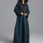 A woman modeling a long dark teal abaya dress with a wide embroidered yoke and matching embroidered cuffs, photographed against a neutral gray studio background.