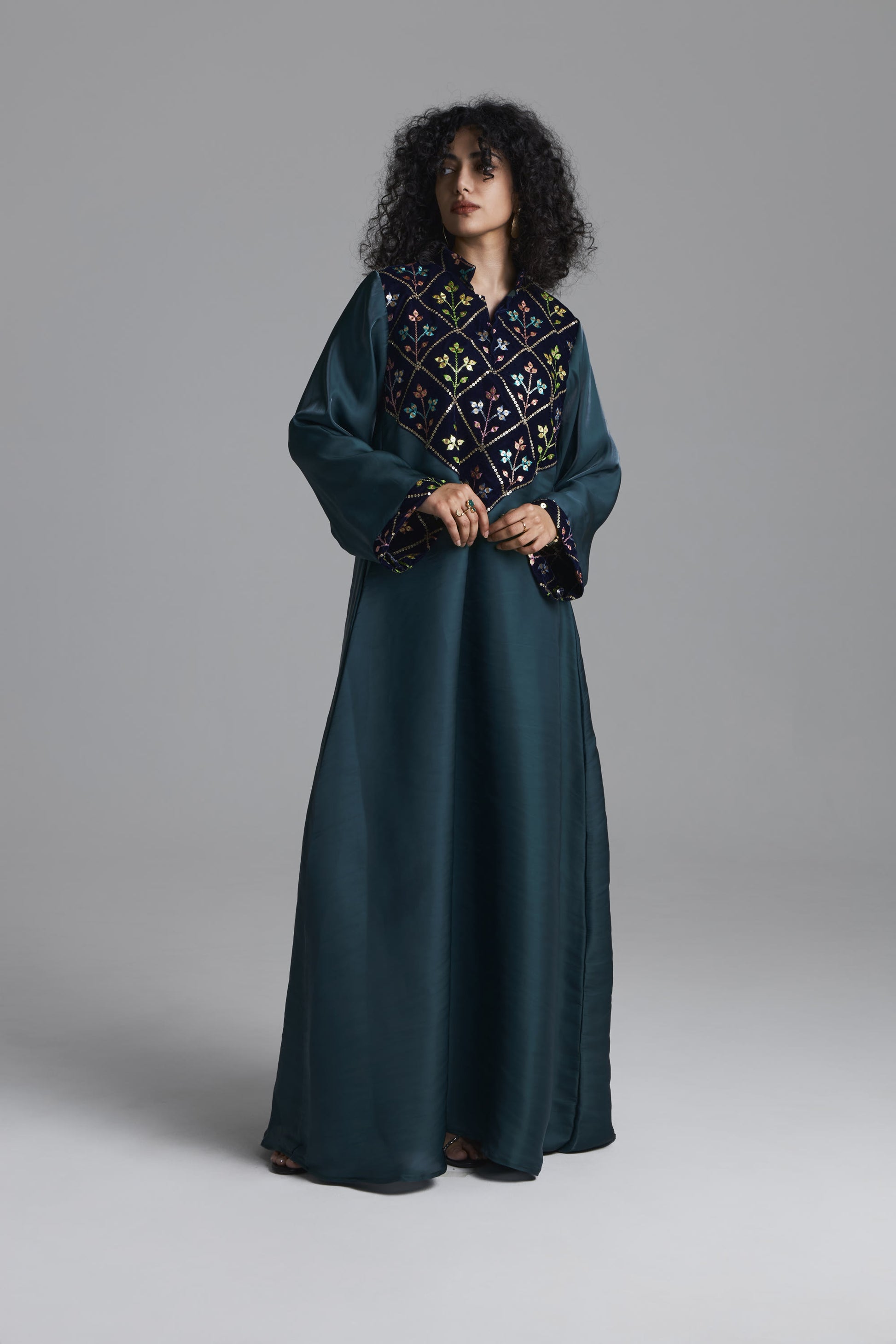 A woman modeling a long dark teal abaya dress with a wide embroidered yoke and matching embroidered cuffs, photographed against a neutral gray studio background.
