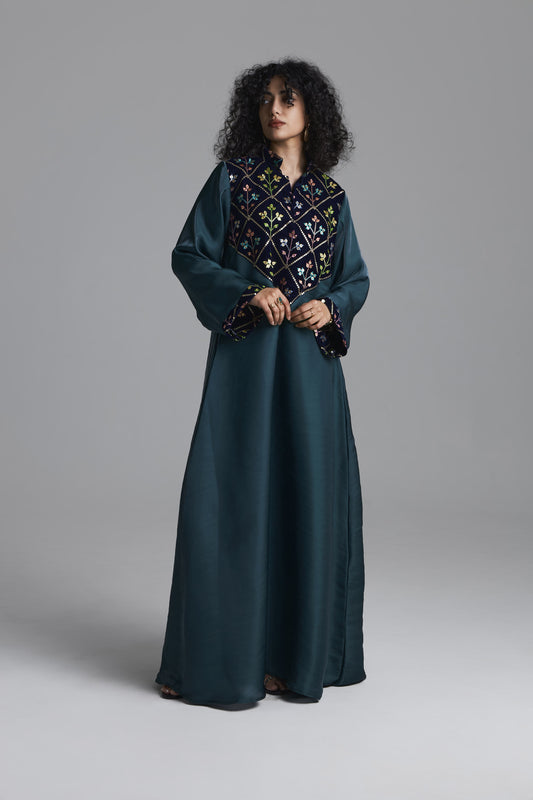 A woman modeling a long dark teal abaya dress with a wide embroidered yoke and matching embroidered cuffs, photographed against a neutral gray studio background.