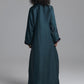 A person viewed from the back wearing a long dark teal satin-like robe or dress with long sleeves and ruffled cuffs, standing against a plain light gray studio background.