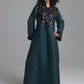 A woman modeling a long dark teal kaftan dress with a large embroidered yoke in black, gold, and white, set against a plain gray studio background.