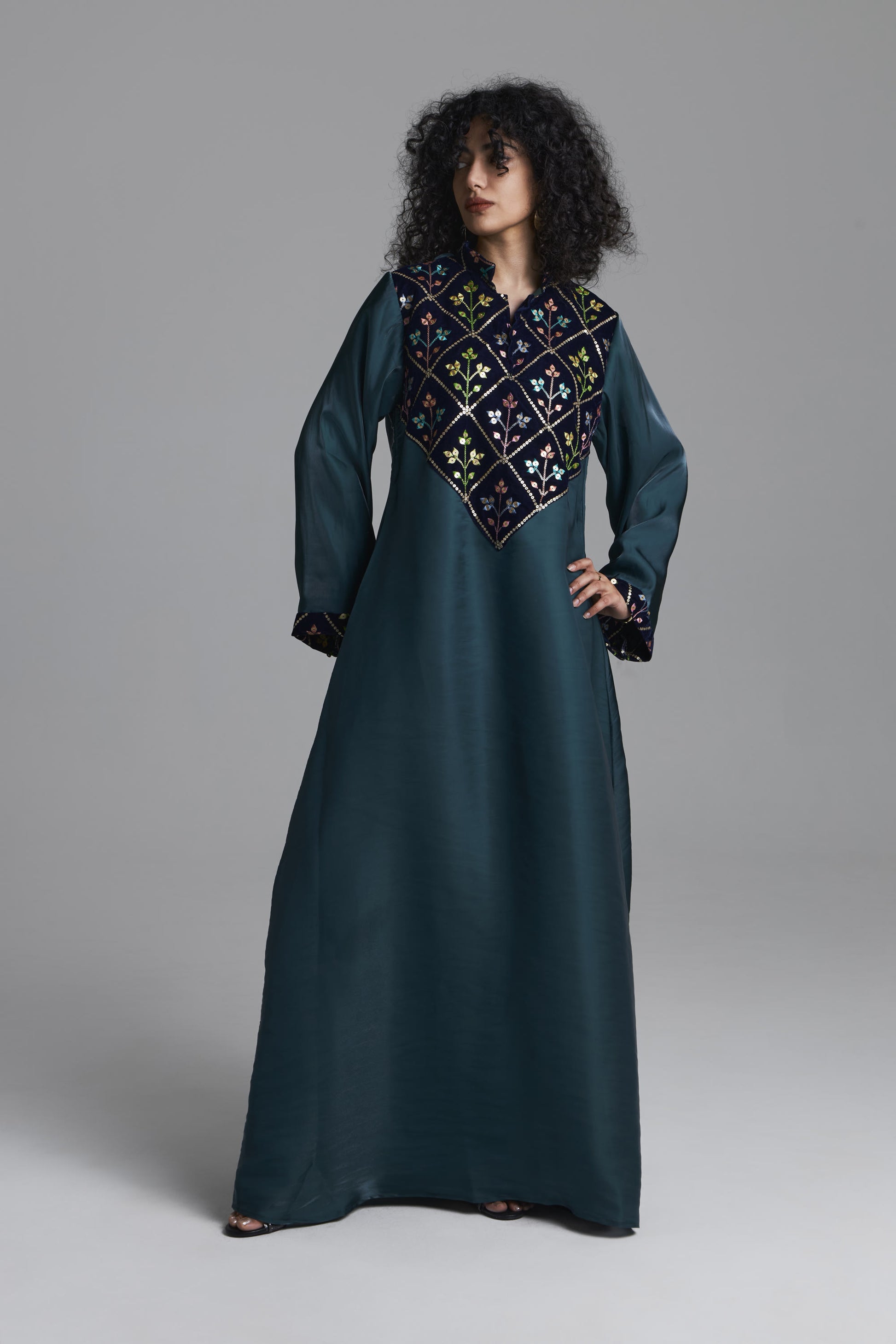 A woman modeling a long dark teal kaftan dress with a large embroidered yoke in black, gold, and white, set against a plain gray studio background.