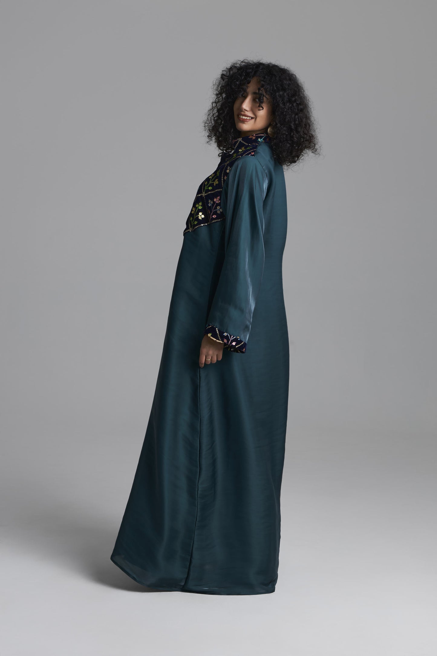 A woman modeling a long, dark teal abaya with intricate multicolor embroidery around the neckline and cuffs, photographed against a neutral gray studio background.