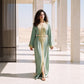 A woman wearing a floor-length sage green kaftan with fine embroidered detailing and a gold belt, standing in a Moroccan-style archway.
